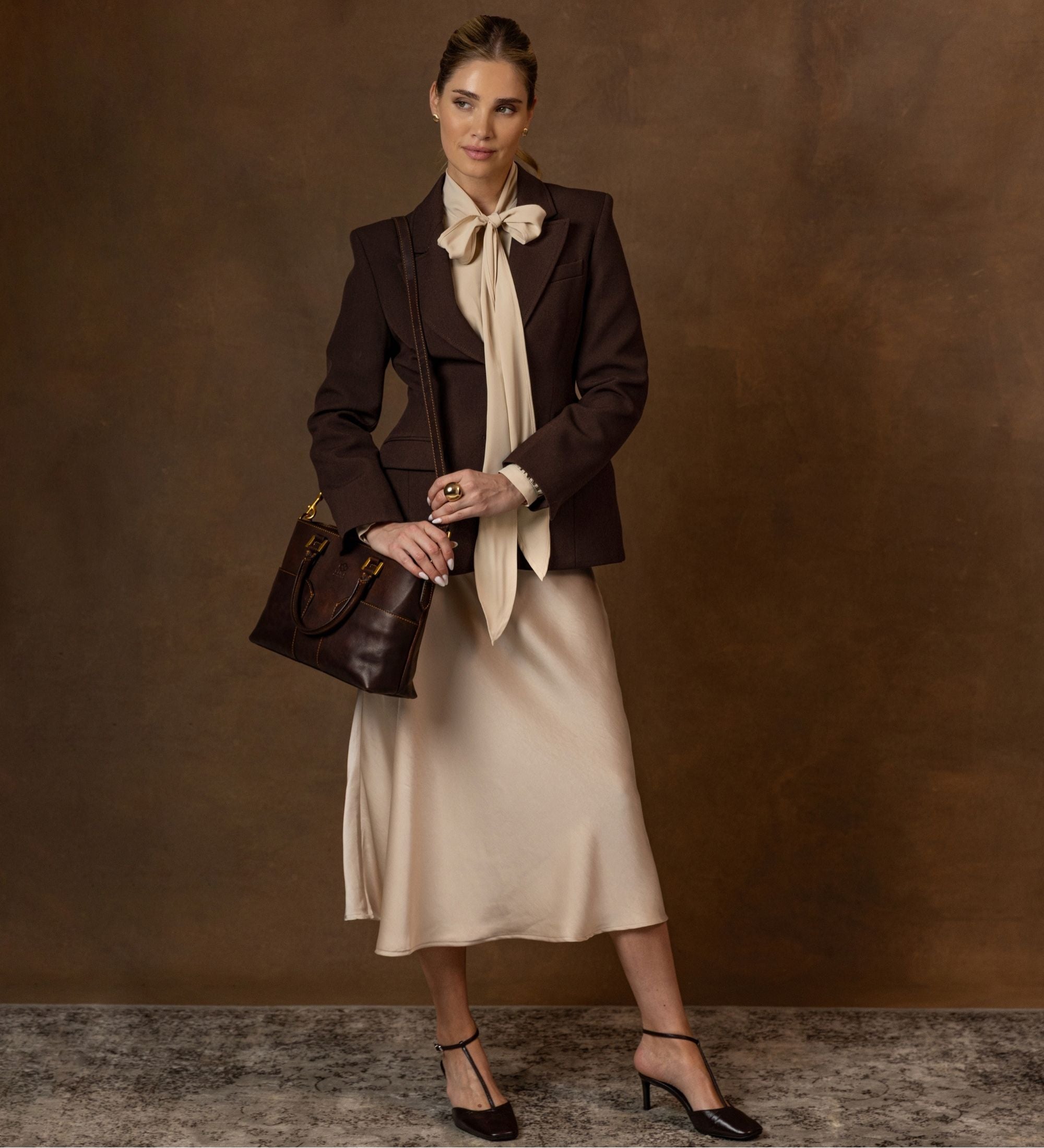 Frau in dunklem Blazer und Bluse mit Schleife steht mit der schokoladenbraunen Camilla Schultertasche in der Hand